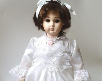 May include: A porcelain doll with brown hair styled with a light blue ribbon bow. The doll wears a white satin dress featuring long sleeves with lace cuffs, a ruffled lace bodice, and a small pink rose accent at the centre of the neckline.