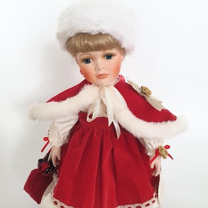 Op de afbeelding: Een porseleinen pop met een rode fluwelen jurk en cape met witte imitatiebont. De pop heeft een witte bontmuts, blond haar en blauwe ogen. Een rode trekkoordtas wordt in de hand van de pop gehouden. De pop is een verzamelobject.