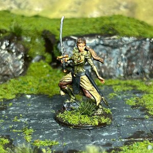 May include: A painted miniature figurine of a female warrior wearing gold and green armor, holding a silver sword. She is standing on a green base.