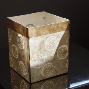 May include: A square-shaped paper lantern featuring a light tan and brown mottled texture with a repeating pattern of white spiral designs. The lantern has a solid brown band along the top edge and an open top, showing a thin metal frame structure inside.