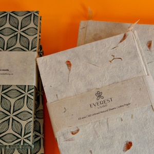 May include: Two stacks of notebooks with a green and white geometric pattern on the cover. The top notebook in the stack on the right has a white cover with brown flecks and the text "EVEREST LIVING A5 size | 180 Unlined Natural Sheets | Lokta Paper".
