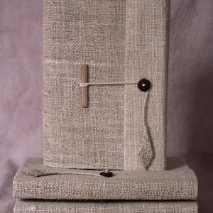 May include: A stack of four linen-covered journals with a wooden button closure and a wooden clothespin on the top journal.