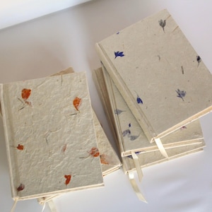 May include: A stack of rectangular journals with textured, off-white covers embedded with small, dried orange and blue flower petals. Each journal features a thin, light-colored ribbon bookmark extending from the bottom edge of the spine.
