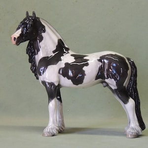 Op de afbeelding: Een keramieken paardenbeeldje in een staande pose. Het paard heeft een zwart-witte vacht met een glanzende afwerking. De manen en staart zijn zwart en de hoeven lichtgrijs. Het paard heeft een roze neus en staat tegen een gedempte groene achtergrond.