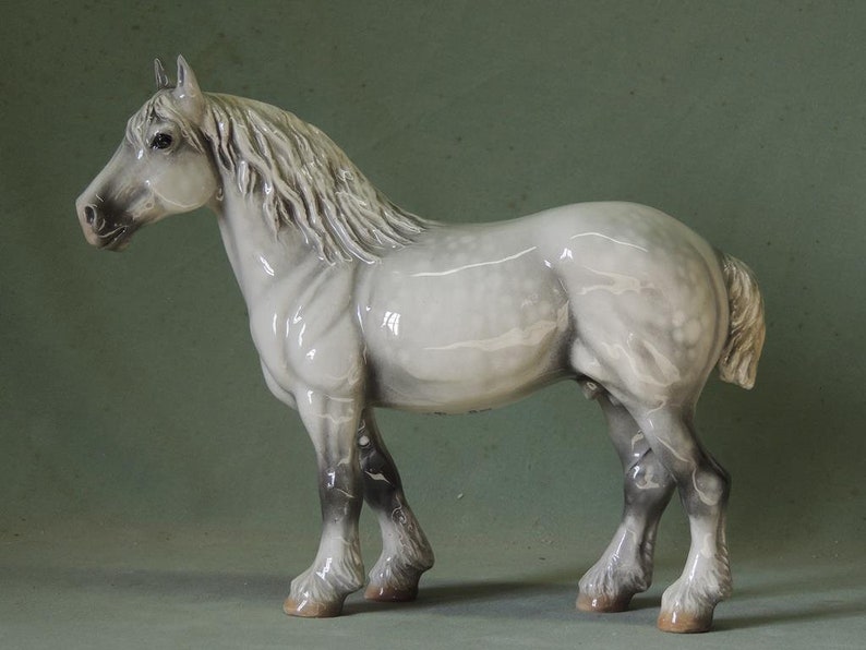 May include: A ceramic horse figurine in a standing pose. The horse is primarily light gray with darker gray accents on its legs and mane. The figurine has a glossy finish and is set against a muted green background.