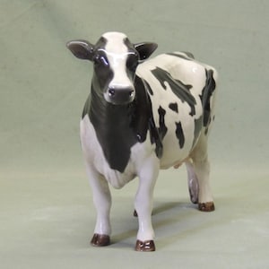 Puede incluir: Una figurita de cerámica negra y blanca de una vaca. La vaca está de pie con las patas separadas y la cabeza levantada. La vaca tiene la nariz negra y las pezuñas negras.