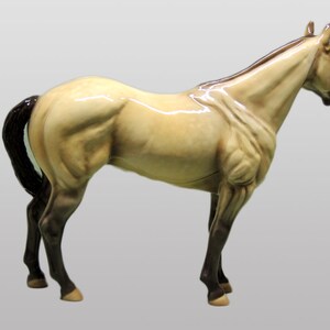May include: A ceramic figurine of a horse in a light brown color with a black mane and tail. The horse is standing on all four legs with its head turned to the side.