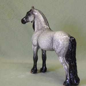 Puede incluir: Una figurita de caballo de porcelana gris y blanca moteada con una crin y cola negras. El caballo est&aacute; de pie con la cabeza girada hacia un lado.