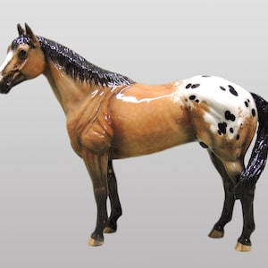 May include: A brown and white spotted horse figurine with a black mane and tail. The figurine is standing on a gray background.