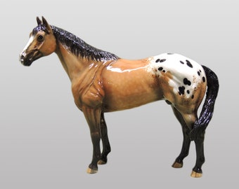 Puede incluir: Una figurita de caballo marrón y blanco con manchas, con una crin y cola negras. La figurita está de pie sobre un fondo gris.