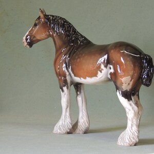 May include: A brown and white ceramic figurine of a Shire horse with a black mane and tail. The horse is standing with its legs spread apart and its head turned to the side.