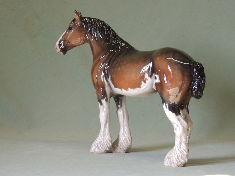 May include: A brown and white ceramic figurine of a Shire horse with a black mane and tail. The horse is standing with its legs spread apart and its head turned to the side.
