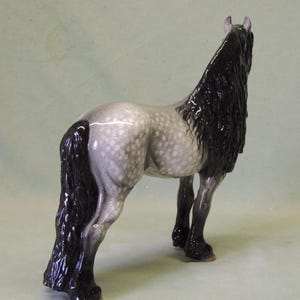 Puede incluir: Una figurita de cer&aacute;mica de un caballo gris y blanco moteado con una larga melena y cola negras. El caballo est&aacute; de pie sobre sus cuatro patas y mira hacia la derecha.