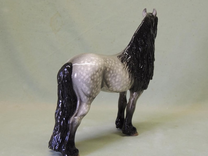Puede incluir: Una figurita de cer&aacute;mica de un caballo gris y blanco moteado con una larga melena y cola negras. El caballo est&aacute; de pie sobre sus cuatro patas y mira hacia la derecha.