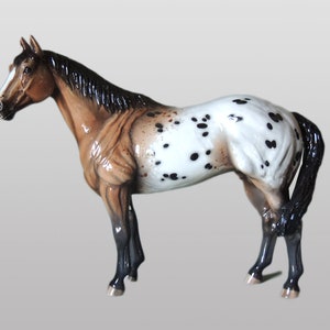 May include: A brown and white spotted horse figurine with a black mane and tail. The figurine is standing on a gray background.