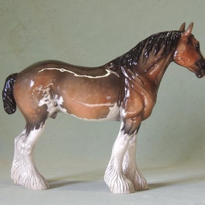May include: A brown and white porcelain figurine of a Shire horse with a black mane and tail. The horse is standing on all four legs with its head turned to the side. The figurine is detailed with white markings on the horse's legs and body.