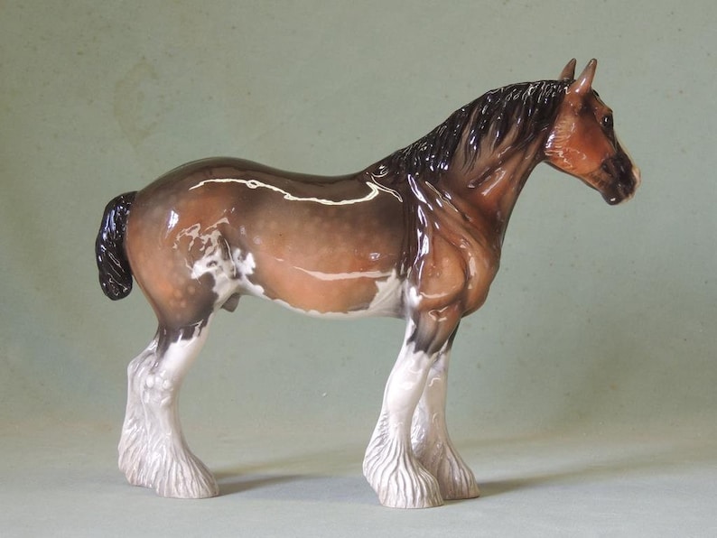 May include: A brown and white porcelain figurine of a Shire horse with a black mane and tail. The horse is standing on all four legs with its head turned to the side. The figurine is detailed with white markings on the horse's legs and body.