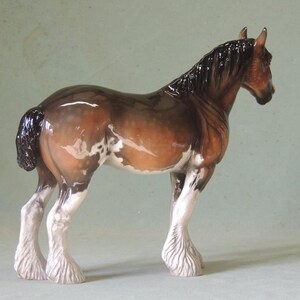 May include: A brown and white porcelain figurine of a Shire horse with a black mane and tail. The horse is standing on all four legs with its head turned to the side.