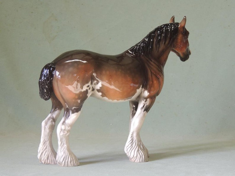 May include: A brown and white porcelain figurine of a Shire horse with a black mane and tail. The horse is standing on all four legs with its head turned to the side.
