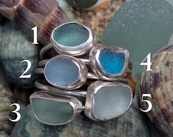 Puede incluir: Una colección de cinco anillos de plata de ley, cada uno con un trozo único de vidrio de mar en tonos azules y verdes. Los anillos se exhiben sobre un lecho de conchas marinas y vidrio de mar, mostrando su belleza natural.