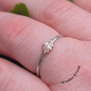 May include: A silver ring featuring a delicate leaf motif in a bronze hue. The leaf is attached to a slender silver band. The ring is displayed on a finger, with the text "Feather & Frost" visible in the lower right corner.
