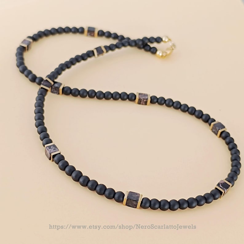 K&ouml;nnte beinhalten: Eine schwarze Perlenkette mit quadratischen, gemusterten Akzenten und goldfarbenen Metalldetails. Die Halskette wird vor einem neutralen Hintergrund pr&auml;sentiert. Die Etsy-Shop-URL ist unten im Bild sichtbar.