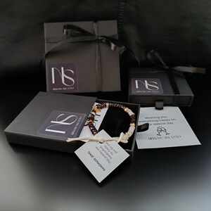 K&ouml;nnte beinhalten: Ein handgefertigtes Armband mit roten und wei&szlig;en Perlen und goldenen Akzenten, pr&auml;sentiert in einer offenen schwarzen Geschenkbox mit schwarzem Band. Die Box enth&auml;lt ein Etikett mit der Aufschrift "HANDMADE JEWEL". Weitere Geschenkboxen mit B&auml;ndern sind ebenfalls sichtbar.