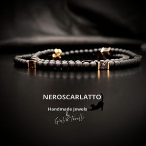 K&ouml;nnte beinhalten: Ein schwarzes Perlenarmband mit goldfarbenen Akzenten und quadratischen, dunkel gef&auml;rbten Steineinlagen. Das Armband wird auf einer reflektierenden schwarzen Oberfl&auml;che pr&auml;sentiert. Der Text "NEROSCARLATTO Handmade Jewels by Giulia Torelli" ist enthalten.
