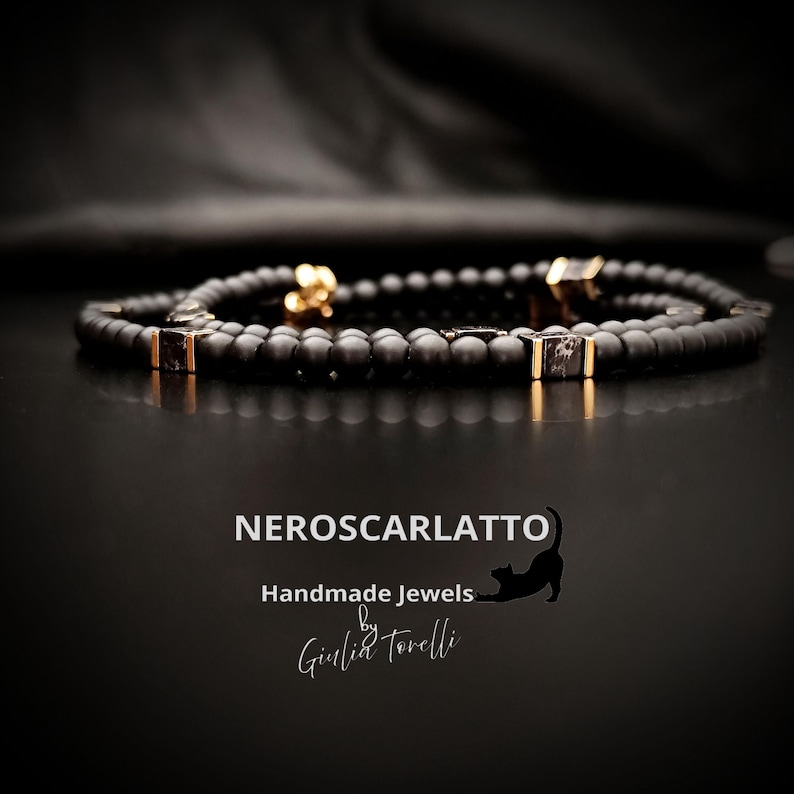 K&ouml;nnte beinhalten: Ein schwarzes Perlenarmband mit goldfarbenen Akzenten und quadratischen, dunkel gef&auml;rbten Steineinlagen. Das Armband wird auf einer reflektierenden schwarzen Oberfl&auml;che pr&auml;sentiert. Der Text "NEROSCARLATTO Handmade Jewels by Giulia Torelli" ist enthalten.