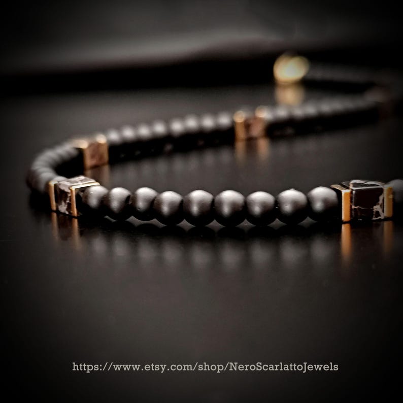 K&ouml;nnte beinhalten: Nahaufnahme einer schwarzen Perlenkette mit goldfarbenen quadratischen Akzenten. Die Halskette besteht aus kleinen, runden Perlen und wird auf einer dunklen Oberfl&auml;che pr&auml;sentiert. Die Etsy-Shop-URL ist unten sichtbar.