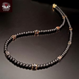 K&ouml;nnte beinhalten: Eine schwarze Perlenkette mit goldfarbenen quadratischen Akzenten. Die Halskette hat kleine, runde schwarze Perlen und mehrere quadratische Perlen mit einem Muster. Handgefertigt in Italien.