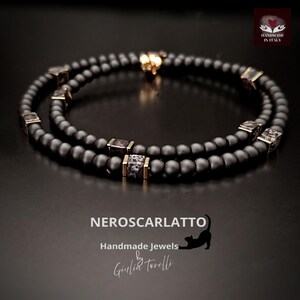 K&ouml;nnte beinhalten: Schwarzes Perlenarmband mit goldfarbenen Akzenten und quadratischen, gemusterten Perlen. Das Armband ist handgefertigt und tr&auml;gt den Text "NEROSCARLATTO Handmade Jewels by Giulia Torelli" auf dunklem Hintergrund.