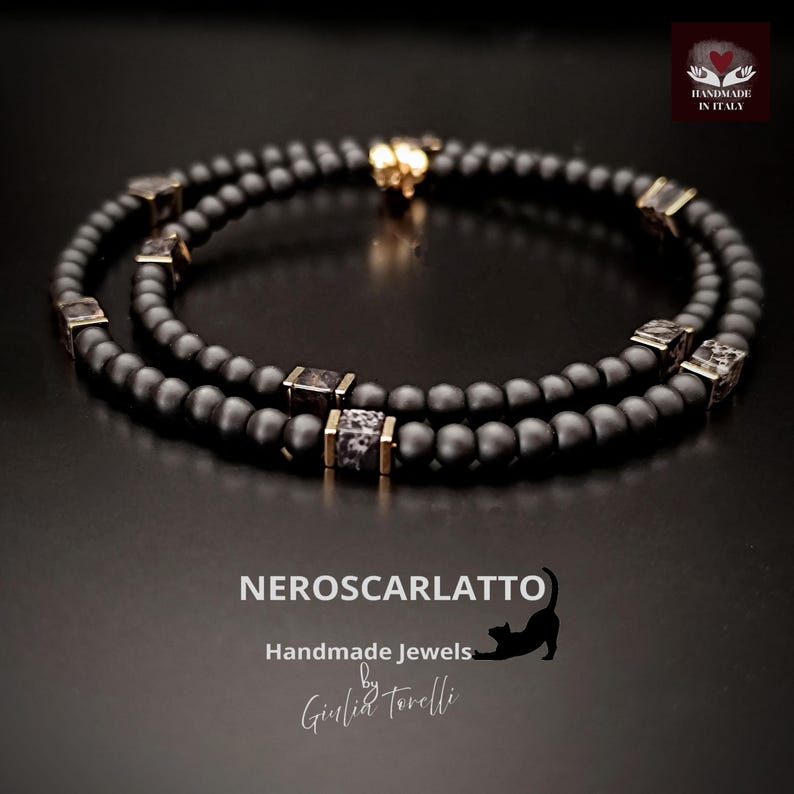 K&ouml;nnte beinhalten: Schwarzes Perlenarmband mit goldfarbenen Akzenten und quadratischen, gemusterten Perlen. Das Armband ist handgefertigt und tr&auml;gt den Text "NEROSCARLATTO Handmade Jewels by Giulia Torelli" auf dunklem Hintergrund.
