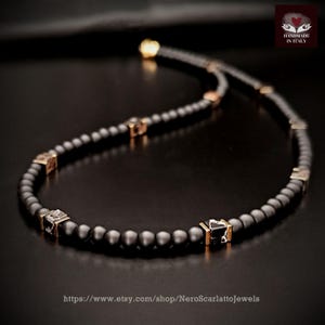 K&ouml;nnte beinhalten: Eine schwarze Perlenkette mit goldfarbenen quadratischen Akzenten und einem goldenen Verschluss. Die Halskette ist handgefertigt in Italien und wird auf dunklem Untergrund pr&auml;sentiert. Die Etsy-Shop-URL ist sichtbar.