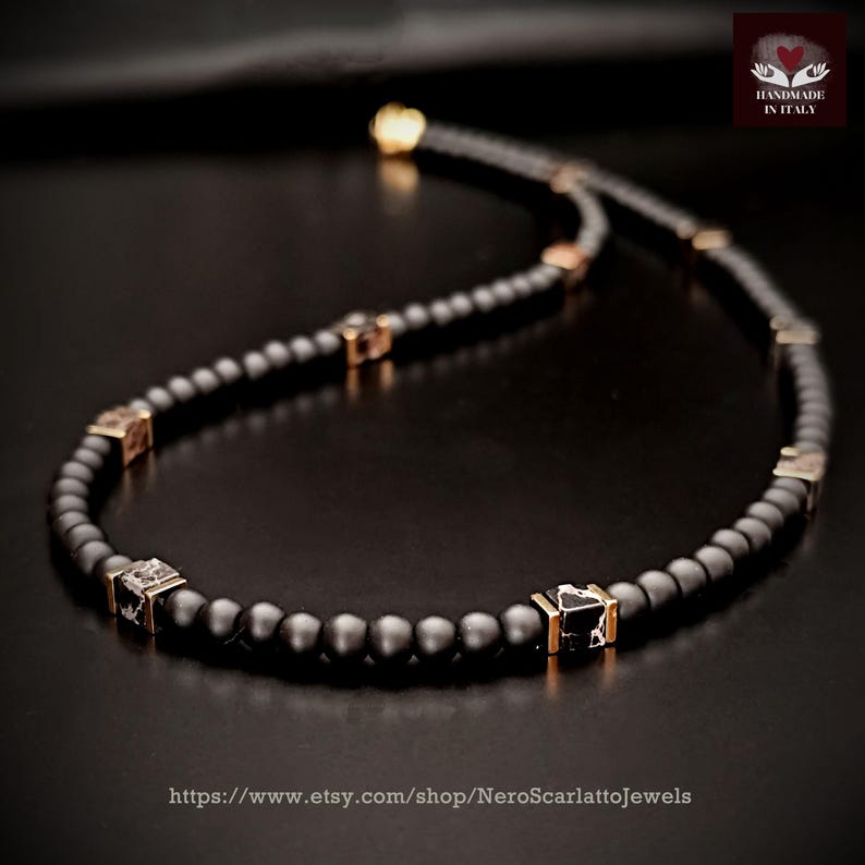 K&ouml;nnte beinhalten: Eine schwarze Perlenkette mit goldfarbenen quadratischen Akzenten und einem goldenen Verschluss. Die Halskette ist handgefertigt in Italien und wird auf dunklem Untergrund pr&auml;sentiert. Die Etsy-Shop-URL ist sichtbar.