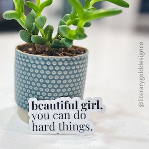 May include: White sticker with black text that reads "beautiful girl, you can do hard things." The sticker is placed on a white surface next to a small potted succulent plant with green leaves.