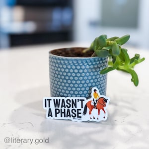 May include: A blue ceramic pot with a textured surface holds a small succulent plant. A white sticker with the text "IT WASN'T A PHASE" and a graphic of a person riding a horse is placed on a white surface in front of the pot.