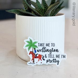 May include: A white sticker with the text "TAKE ME TO Wellington & TELL ME I'M pretty". The sticker features a palm tree, a person on a horse, and the hashtag #literarygold.