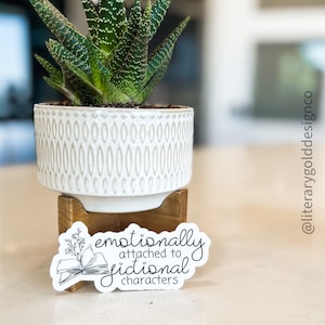 May include: A black and white sticker with the text "emotionally attached to fictional characters" and a drawing of an open book with a flower sprig on top. The sticker is on a white surface next to a potted succulent.