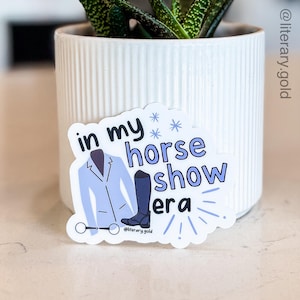 May include: A sticker with the text "in my horse show era" in blue and white. The sticker features a light blue riding jacket, a dark blue riding boot, and a pair of riding glasses. The sticker is placed in front of a white ceramic pot with a green plant.