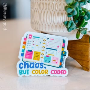 May include: A die-cut sticker featuring an open planner with colorful sticky notes, checklists, and grid pages. The text below reads "chaos, BUT COLOR CODED" in blue, yellow, and pink lettering. The design includes various icons like hearts and stars.