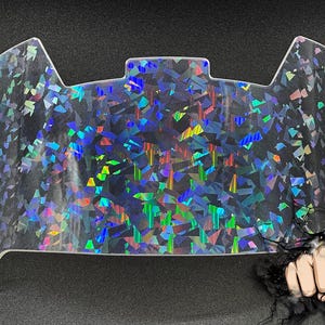 May include: A clear football helmet visor featuring a holographic, multi-colored shattered glass pattern that reflects light in shades of blue, green, and yellow. The visor has a curved top edge with a central tab and mounting holes on the sides.