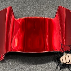 May include: A glossy, red automotive part with a curved design. The object has a reflective surface and appears to be made of a hard material. The shape suggests it could be a component of a vehicle's exterior, possibly a trim piece or a light housing.