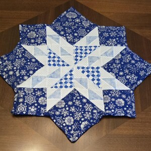 May include: A blue and white quilted star-shaped placemat. The placemat features a central star design with white and blue geometric patterns. The outer edges are a blue fabric with a silver snowflake pattern.