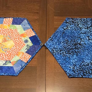 May include: Four handmade hexagon-shaped fabric coasters. Two are solid blue with a floral pattern, and two feature a colorful patchwork design with orange, yellow, and green accents. The coasters are arranged on a dark wooden surface.