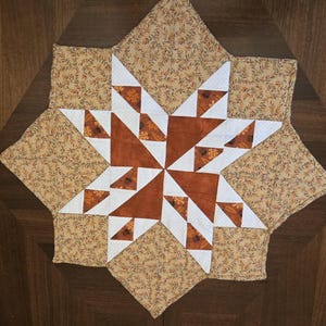 May include: A star-shaped quilted placemat with a central star design in shades of brown, white, and tan. The star is surrounded by a tan fabric with a leaf pattern. The placemat is placed on a dark wood surface.
