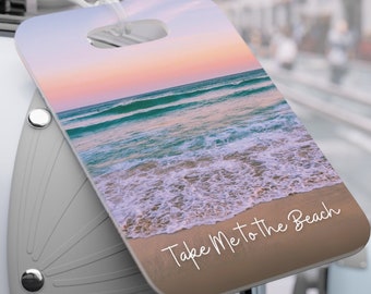 以下が含まれることがあります： 夕暮れのビーチの風景と「Take Me to the Beach」というテキストが書かれた荷物タグです。