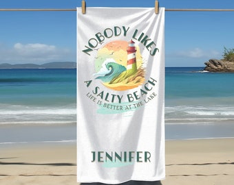 以下が含まれることがあります： 白いビーチタオルに、灯台、波、そして「Nobody Likes A Salty Beach Life Is Better At The Lake」というテキストのカラフルなイラストが描かれています。タオルには「Jennifer」という名前も印刷されています。