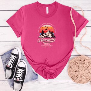 May include: A fuchsia t-shirt with rolled-up sleeves featuring a mountain and sunset graphic. The text reads "The Smoky Mountains are Calling, Adventure Awaits, Johnson Family Vacation 2024." Black canvas sneakers and denim shorts are next to the shirt.