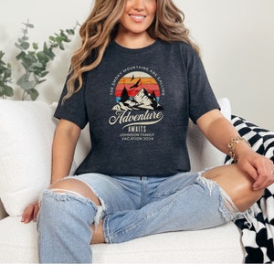 May include: Dark gray t-shirt featuring a mountain and sunset graphic with the text "The Smoky Mountains Are Calling, Adventure Awaits, Johnson Family Vacation 2024". The shirt is a crew neck and is made of a soft material.